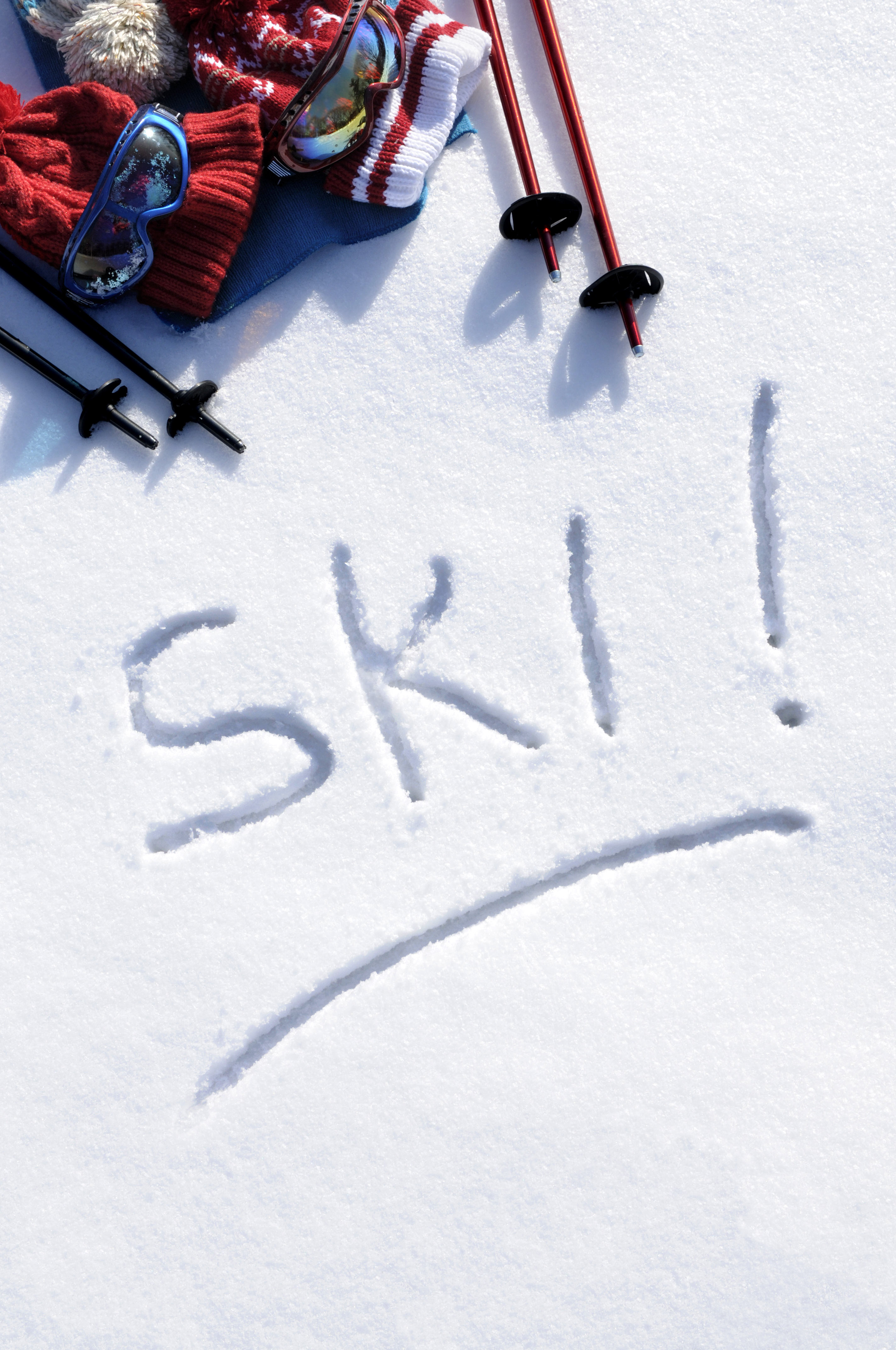 Ski gear essentials including goggles, gloves, hat and poles laid on fresh snow with &ldquo;ski&rdquo; written in the snow